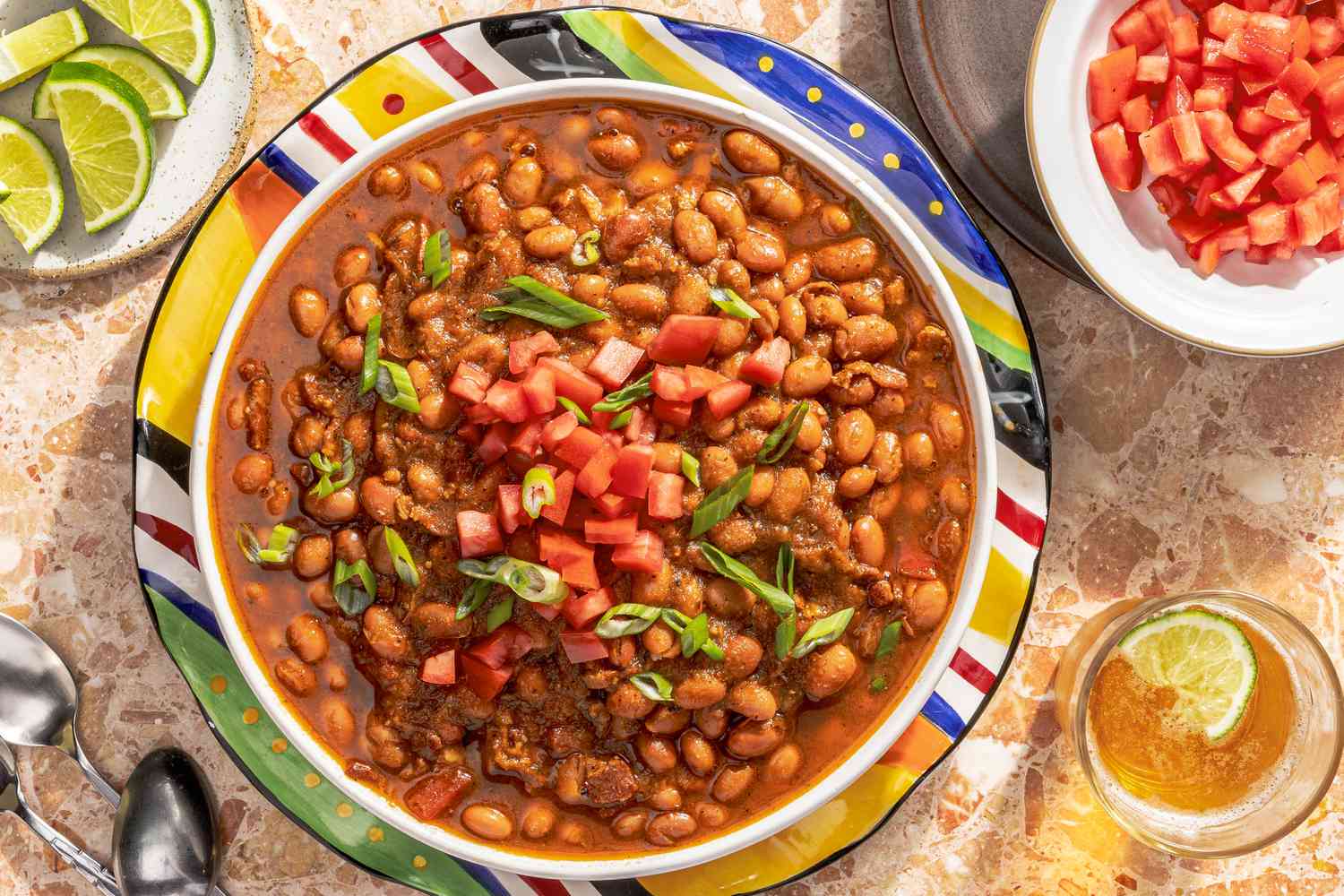 borracho beans (frijoles beans) in a bowl at a table setting with a bowl of diced tomatoes, small plate with lime wedges, a glass of beer, and utensils
