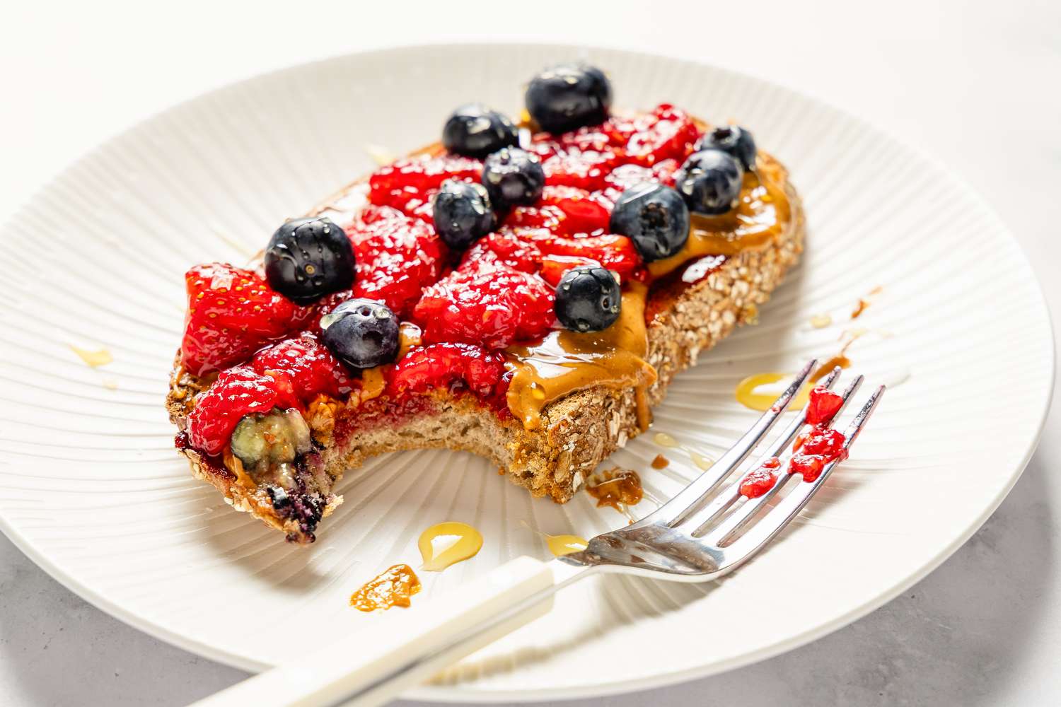 A piece of toast with peanut butter, strawberries, and blueberries, partially eaten, on a white plate with a fork nearby