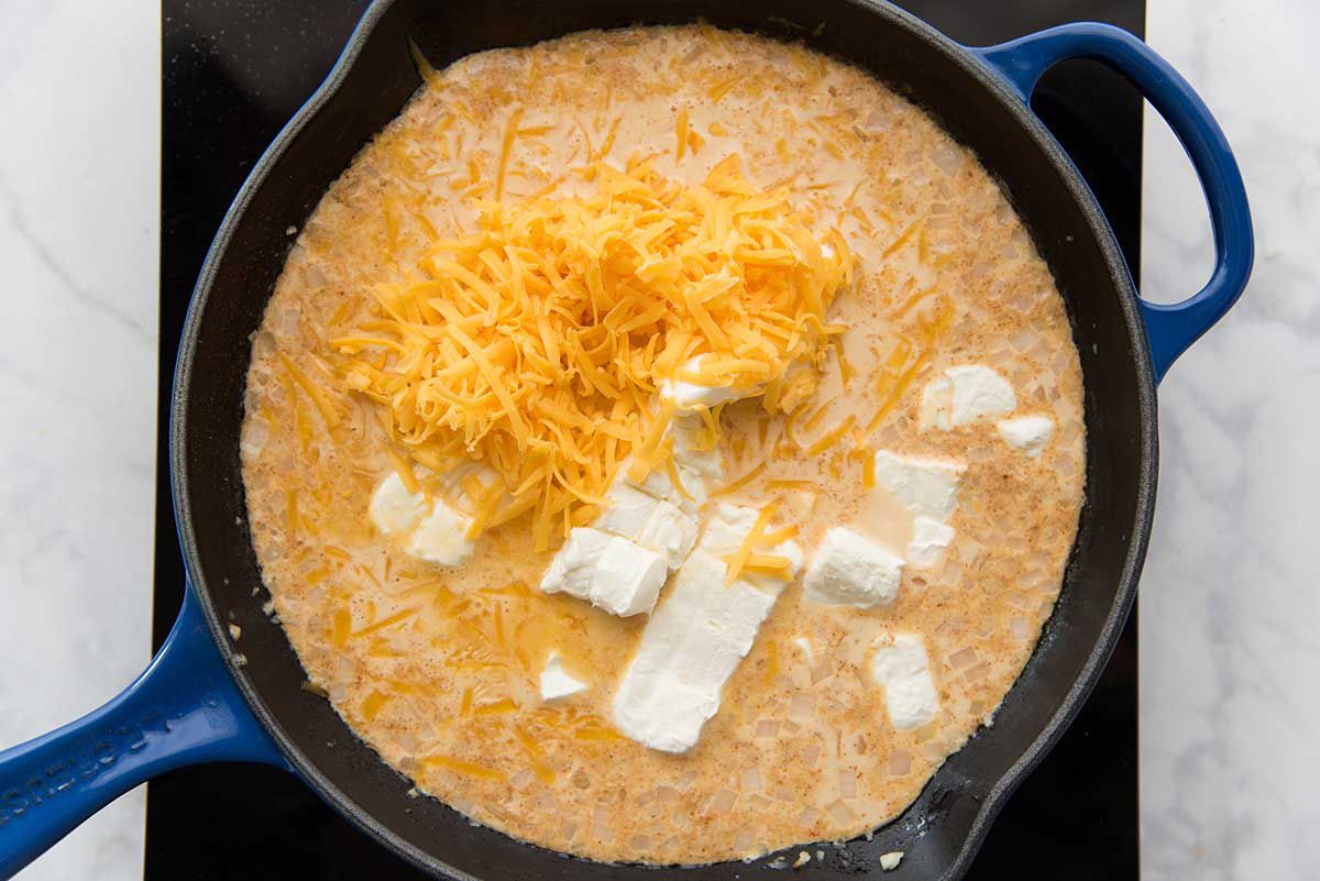 A black cast iron skillet will spices, milk and cheese being stirred together with a wooden spoon for gameday queso