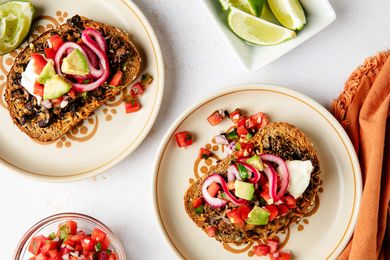 Plates of black bean toast topped with avocado pickled onions and garnishes accompanied by lime wedges and a bowl of salsa