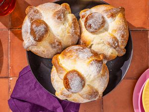 Pan de muerto in a bowl next to a stack of plates, a purple kitchen towel, and glasses of water, all on a brick surface