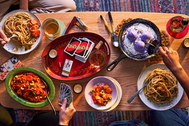 Coffee table filled with food including pasta, carrot salad, and ube skillet cookie, Hands are digging into the food and playing uno