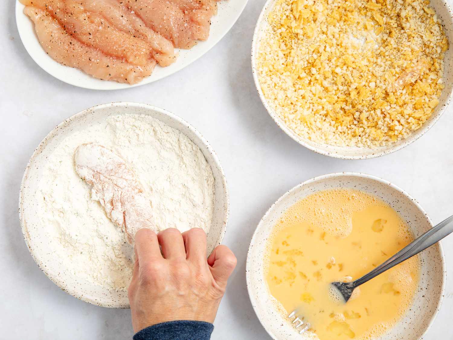 hand putting chicken tenders in a bowl of flour; bowl of egg mixture, breadcrumb mixture, and a plate of raw seasoned chicken tenders