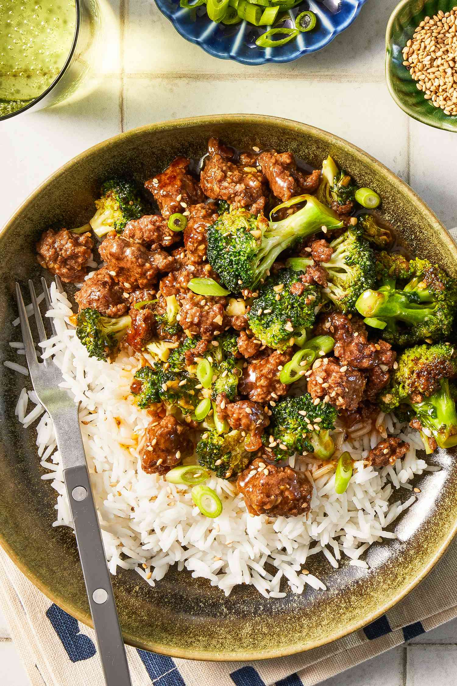 Ground beef and broccoli stir fry over rice in a bowl at a table setting with a glass of water, a bowl of sliced scallions, and a bowl of sesame seeds