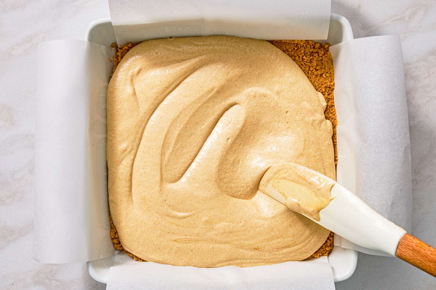 overhead view of square baking dish with cookie crumb crust and peanut butter mixture being spread on top for Chocolate-Peanut Butter Delight