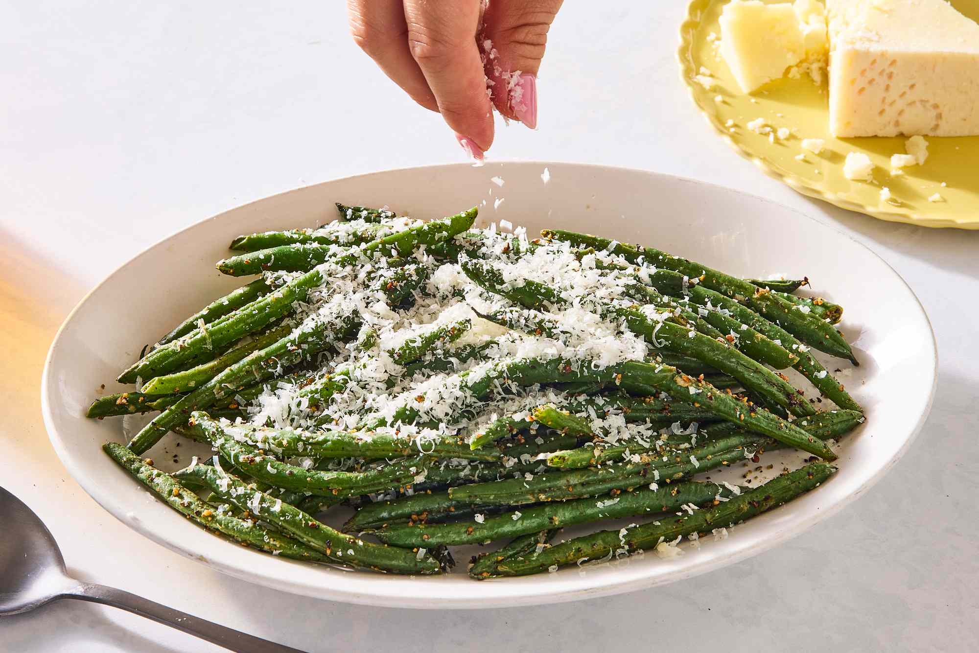 A plate of green beans topped with grated cheese a hand sprinkling cheese over them