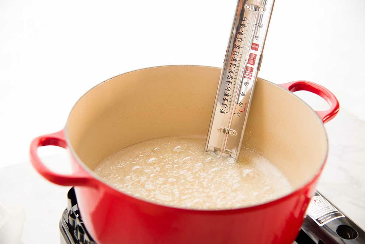 Side view of a dutch oven simmering on the stove with light colored sugar simmering with a candy thermometer in the pot to make Classic Caramel Candies.