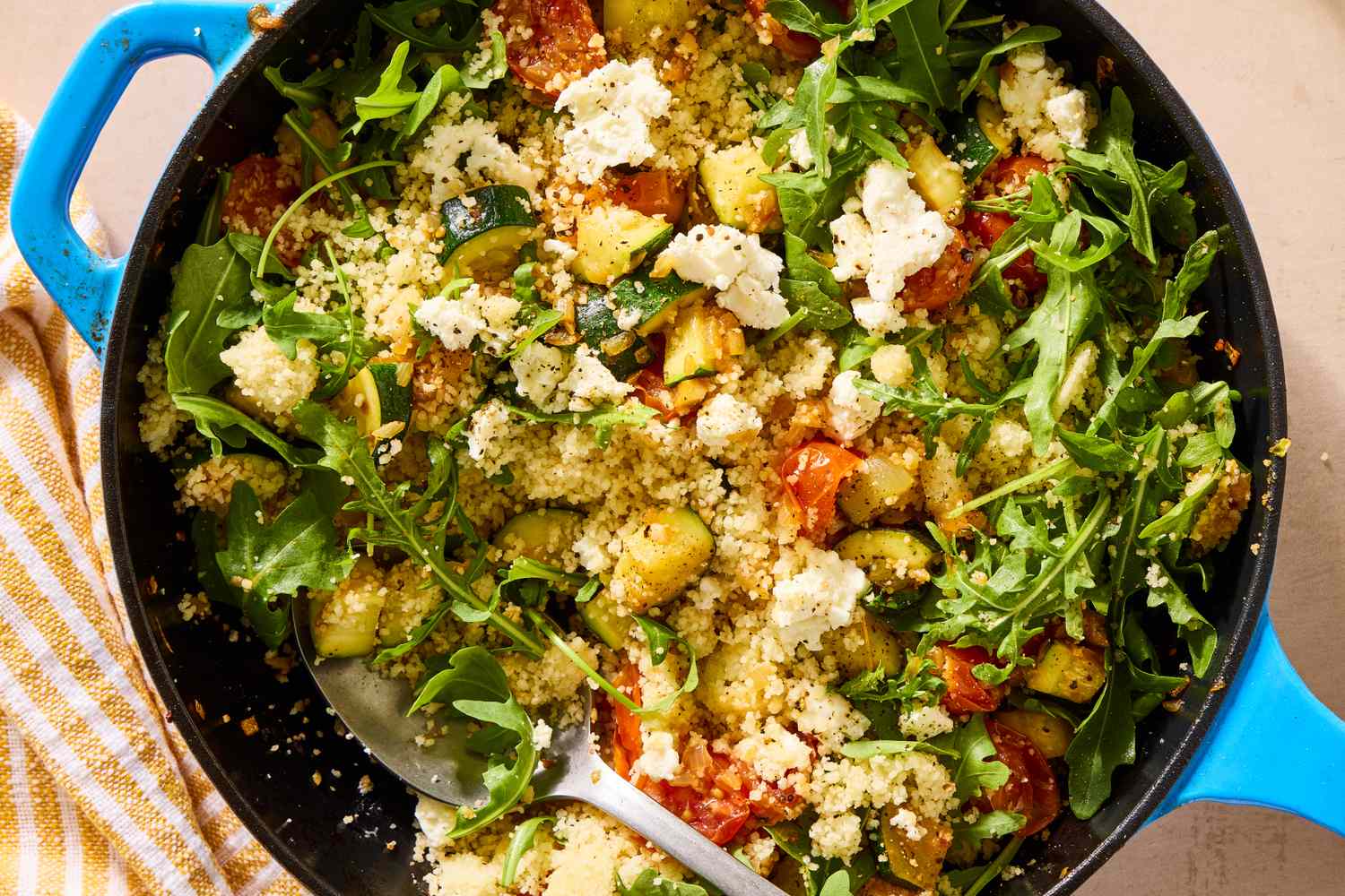 A skillet containing couscous, arugula, roasted vegetables, and cheese crumbles, with a serving spoon