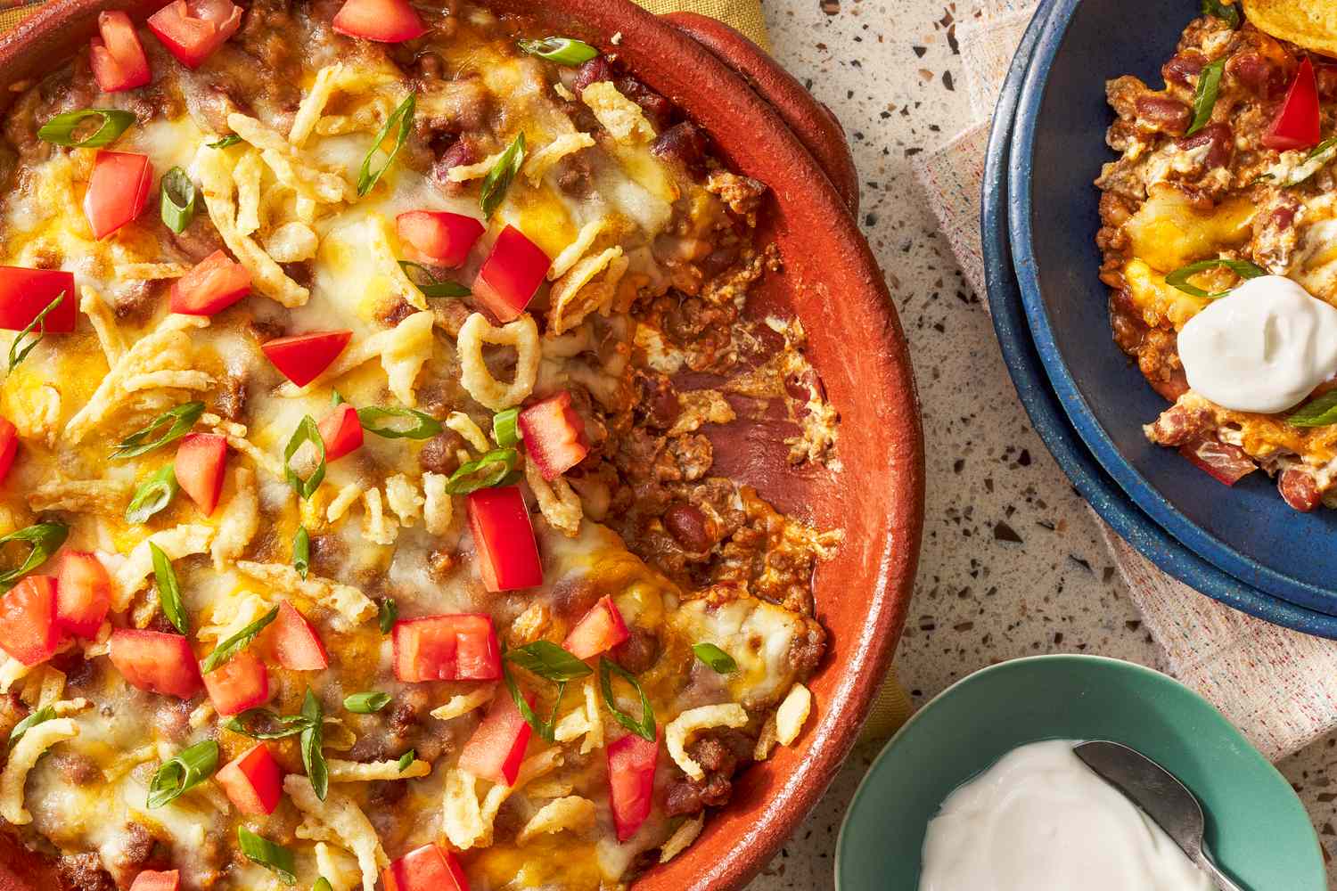 Baked dip in a casserole dish garnished with diced tomatoes and green onions next to a plate with a serving and a dollop of sour cream