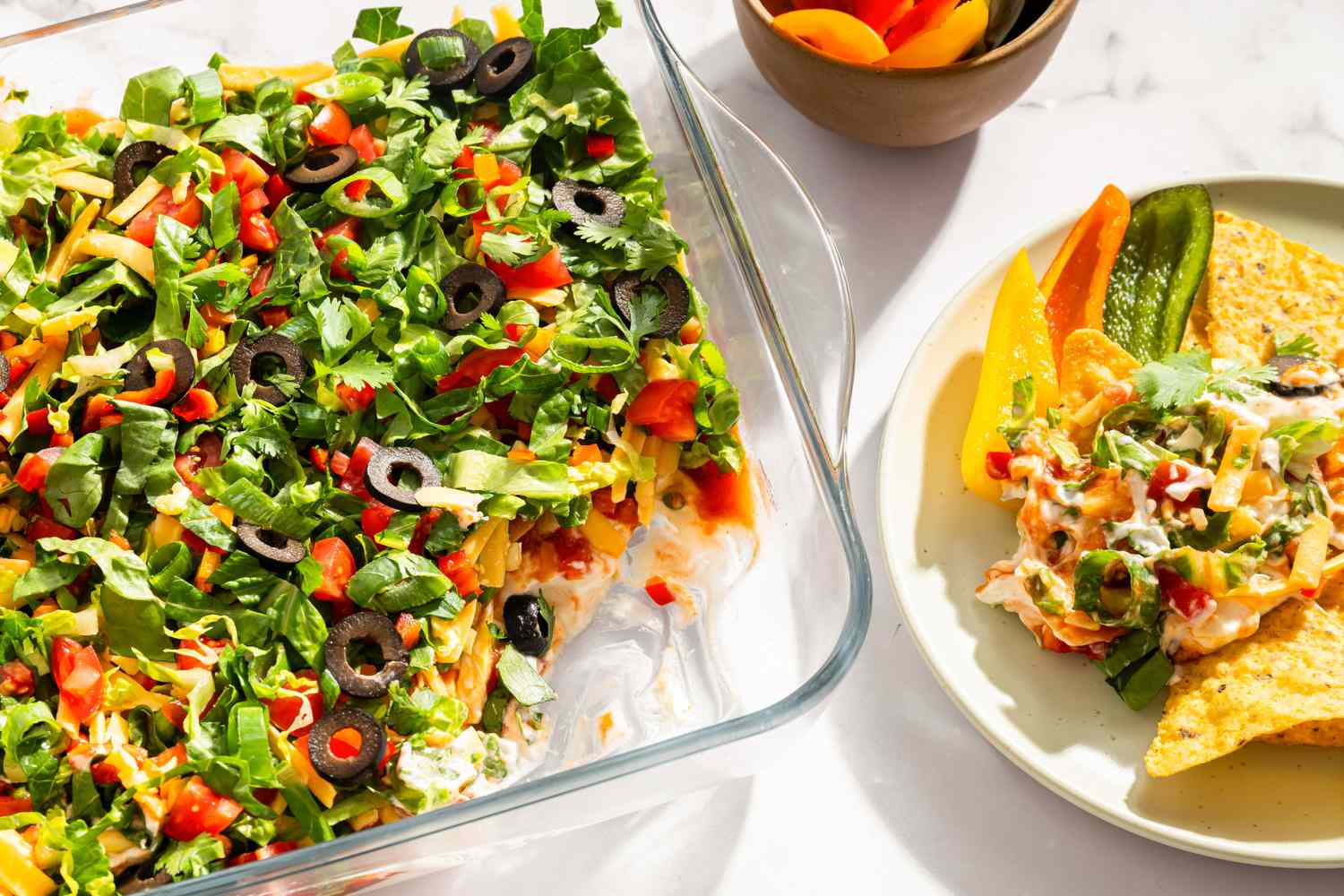 A glass dish containing layered nacho dip topped with ingredients such as olives herbs and vegetables served with tortilla chips on a plate