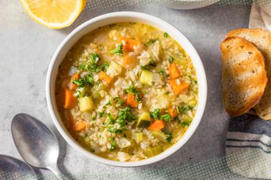 Bowl of rice and vegetable soup, and in the surroundings, slices of toast on a white and blue kitchen towel, a halved lemon, and utensils on the counter