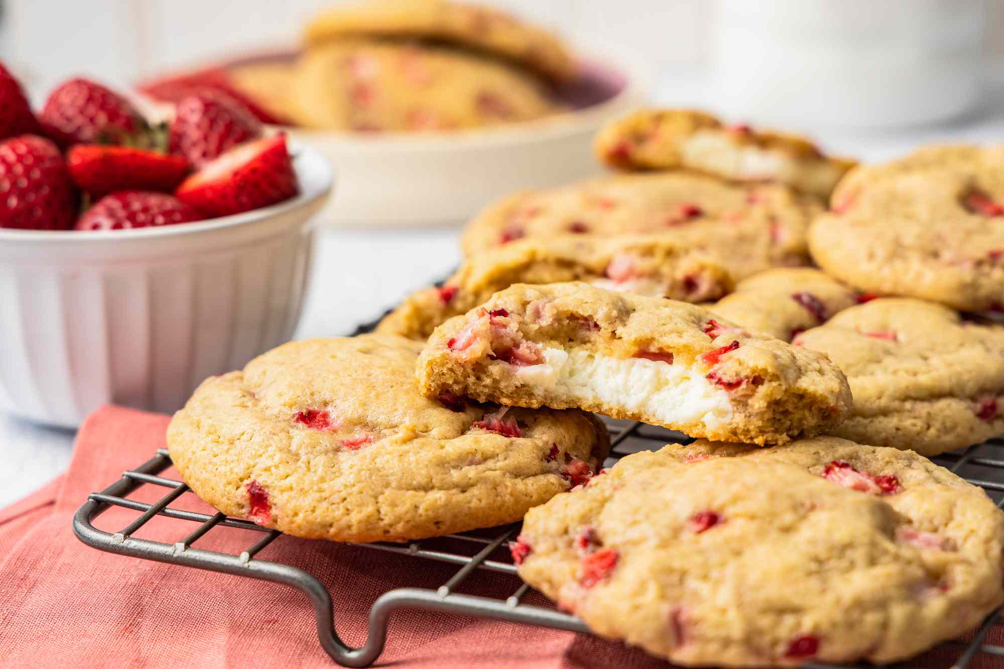 Strawberry Cheesecake Cookies on a Cooling Rack With Some Cut in Half, and in the Surroundings, a Bowl of Whole Strawberries and a Plate With More Cookies, All on a Pink Table Napkin