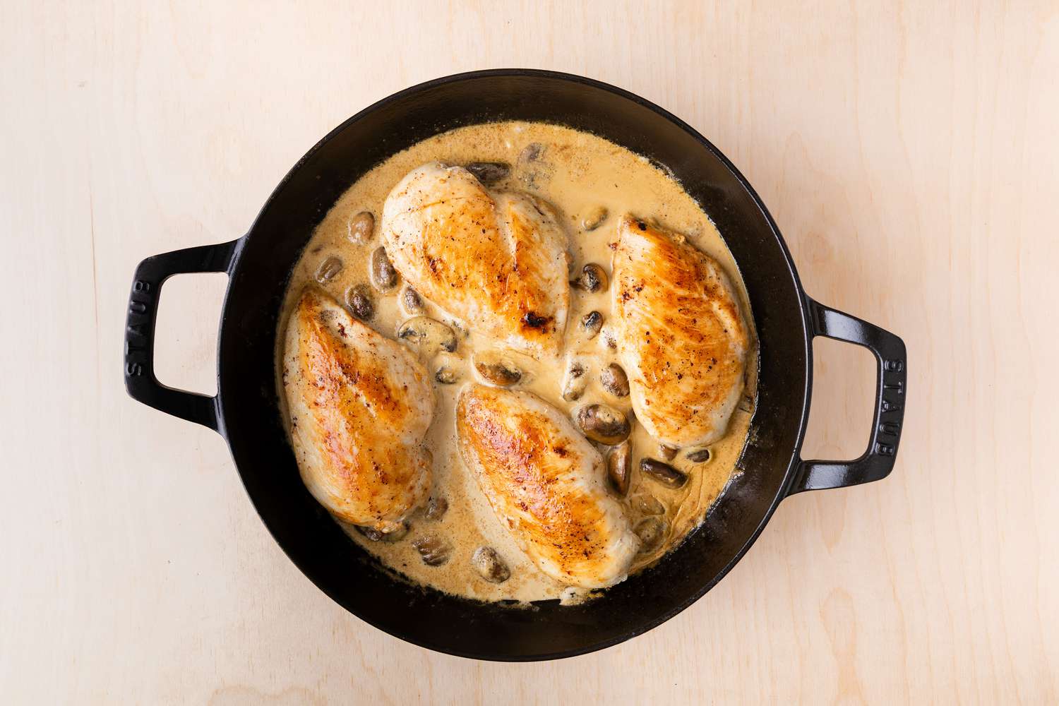 overhead view of cream sauce, thickened with browned chicken breasts added to the top in a cast iron skillet for browned chicken breasts being added to cream sauce with mushrooms in large cast iron skillet for halved button mushrooms browning in a large cast iron skillet for Chicken Supreme