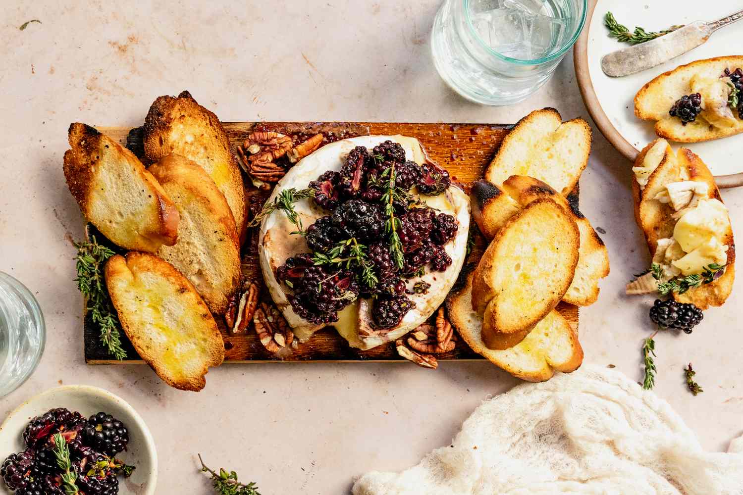 Indoor Table Setting: Cedar Plank Grilled Brie Topped With Black Berries and Sprigs of Thyme, Slices of Baguette on the Plank Next to the Brie, and on the Table, Two Glasses of Water, Bowl of Blackberries, a Plate With Slices of Baguette Topped With Brie and Blackberries, and a Kitchen Linen