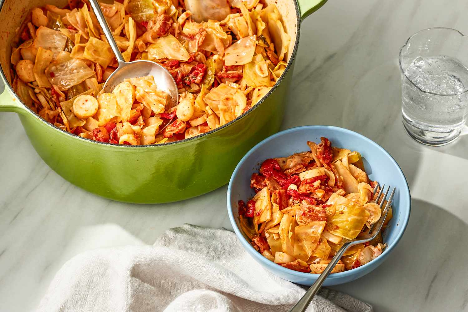 Overhead view of a green dutch oven of swamp cabbage with a metal serving spoon and a small blue bowl and fork of cabbage