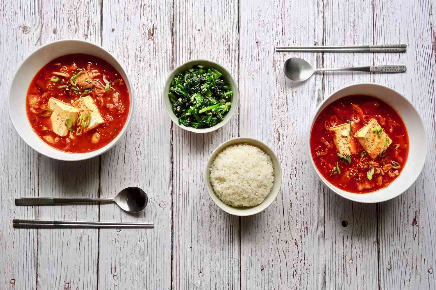 Korean kimchi and pork belly stew in two bowls with utensils, rice, and herbs set between them.