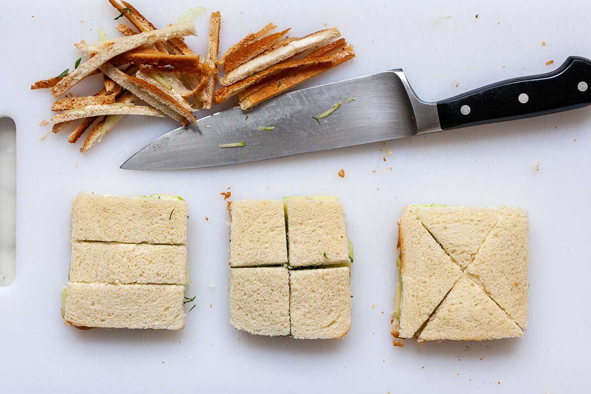 Showing different ways to slice cucumber tea sandwiches. A chef's knife is above three sandwiches and the crusts are cut off.