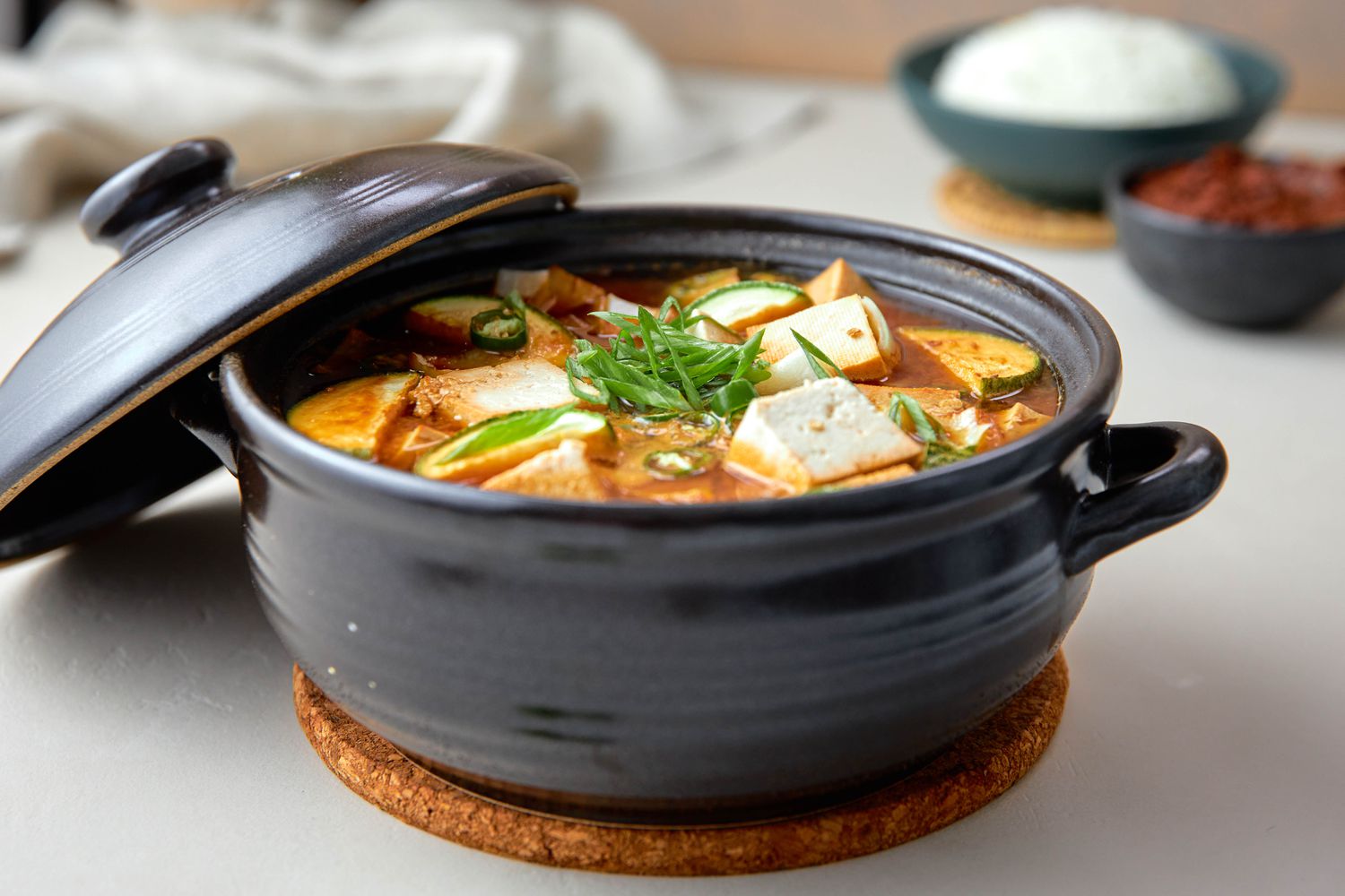 Doenjang Jjigae (Soybean Stew) in a Clay Pot on the Counter, and in the Surroundings, a Bowl of Rice, a Bowl of Banchan, and a White Kitchen Towel
