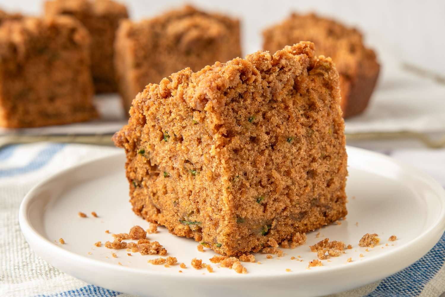 A slice of zucchini coffee cake on a white plate with a crumb topping and background showing additional slices on a cooling rack