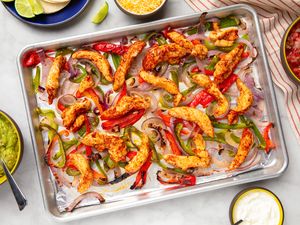 Sheet pan with cooked chicken strips and assorted vegetables surrounded by small bowls of guacamole pico de gallo and condiments