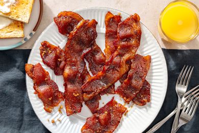 Overhead view of millionaire's bacon on a white plate next to a glass of orange juice, a plate of toast and three forks