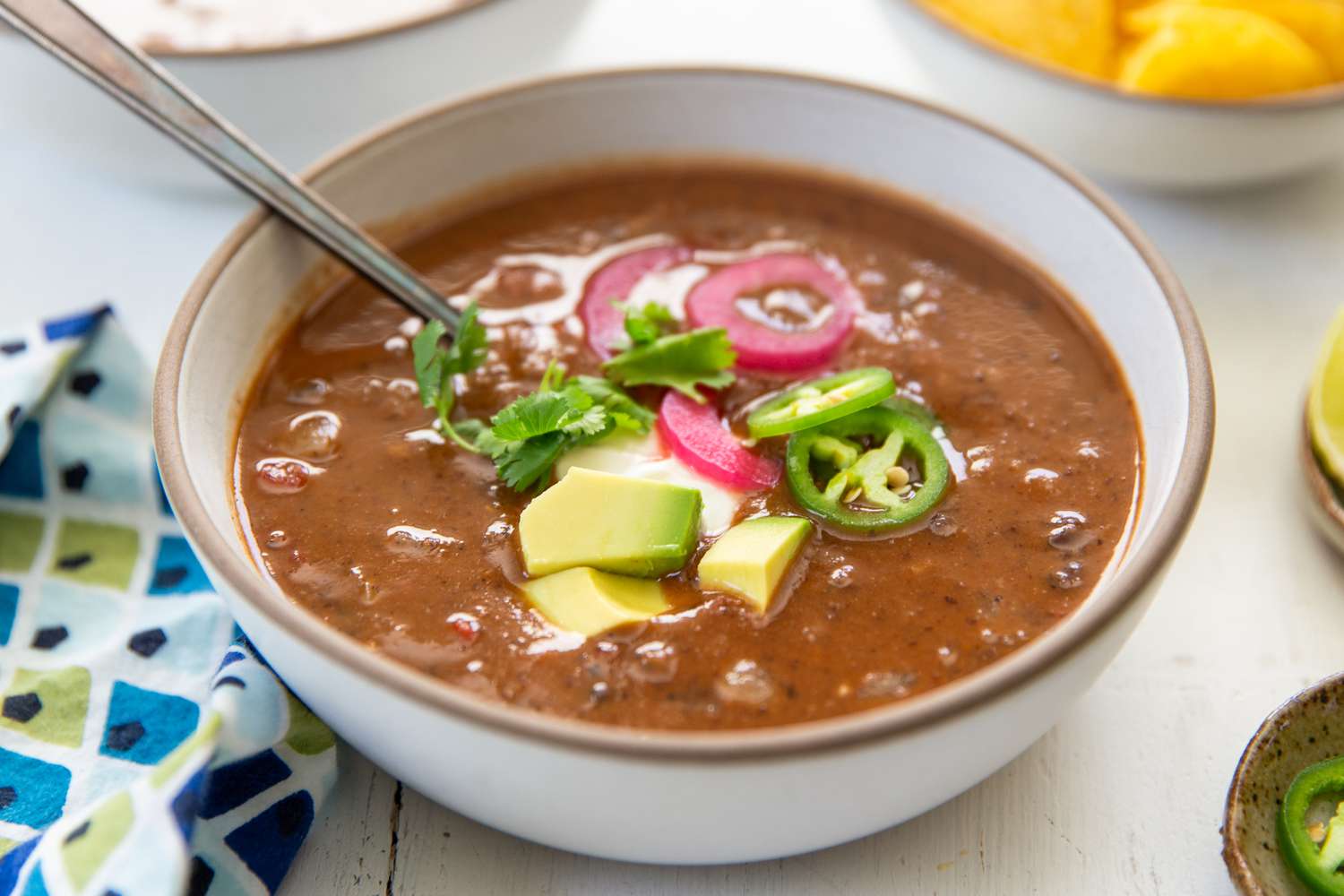 A bowl of black bean soup, topped with avocado cubes, jalapeño slices, pickled onion and and cilantro