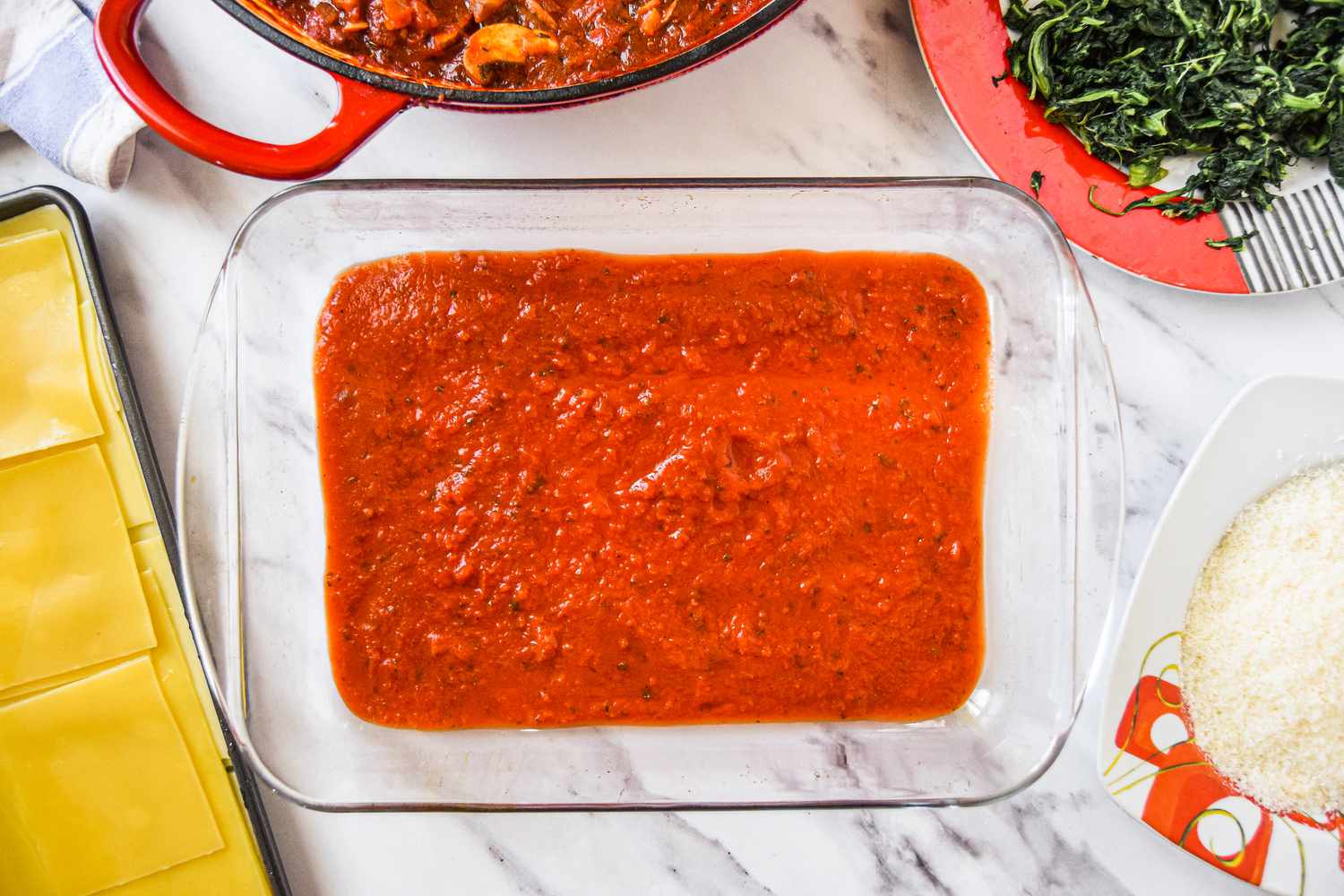A layer of Sauce in a Casserole Dish Surrounded by a Tray with Lasagna Noodles, a Bowl with Grated Parmesan Cheese, a Bowl of Spinach, and a Dutch Oven of Sauce