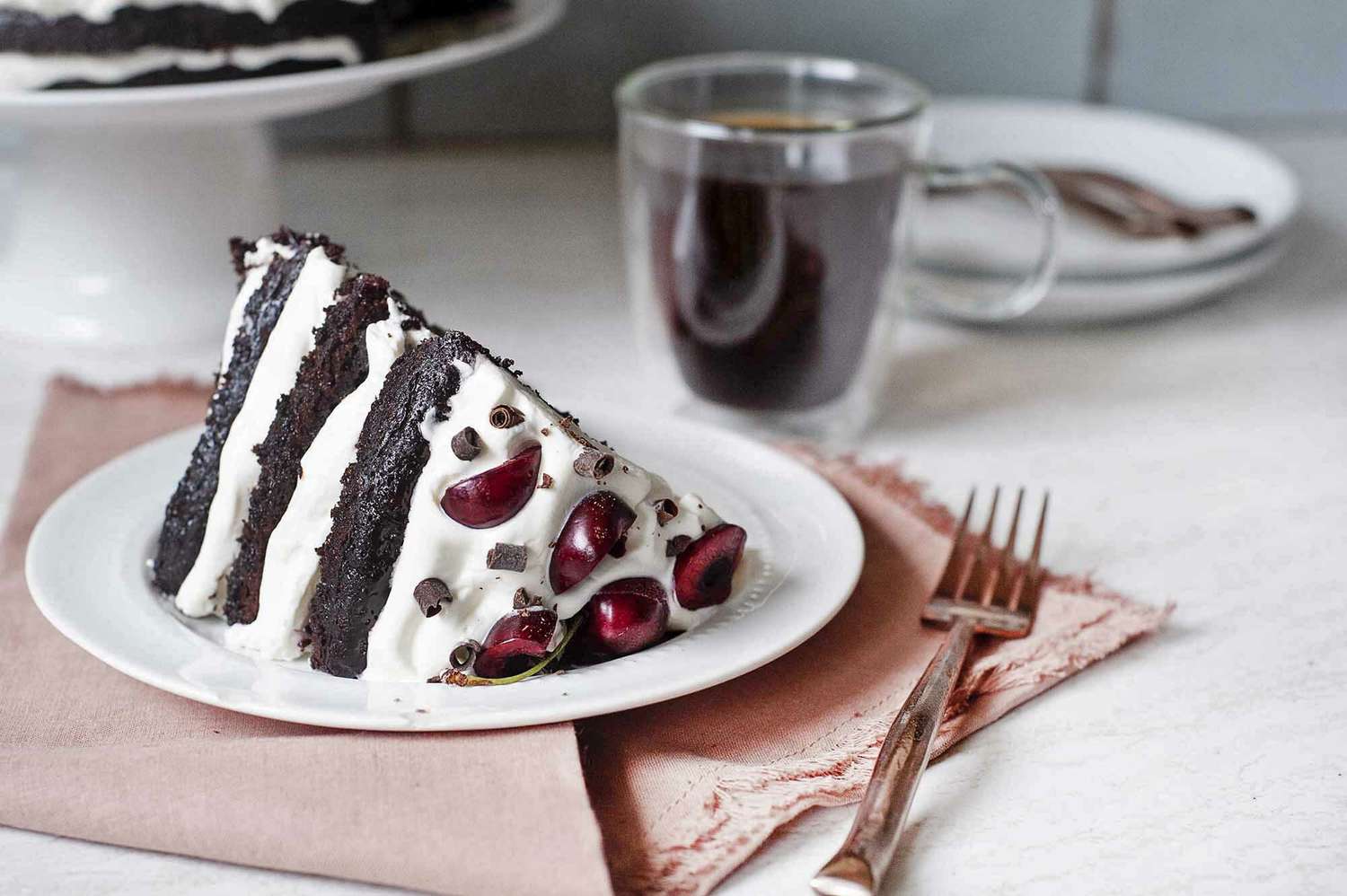 Authentic Black Forest Cake on a white plate with a cup of tea set behind.