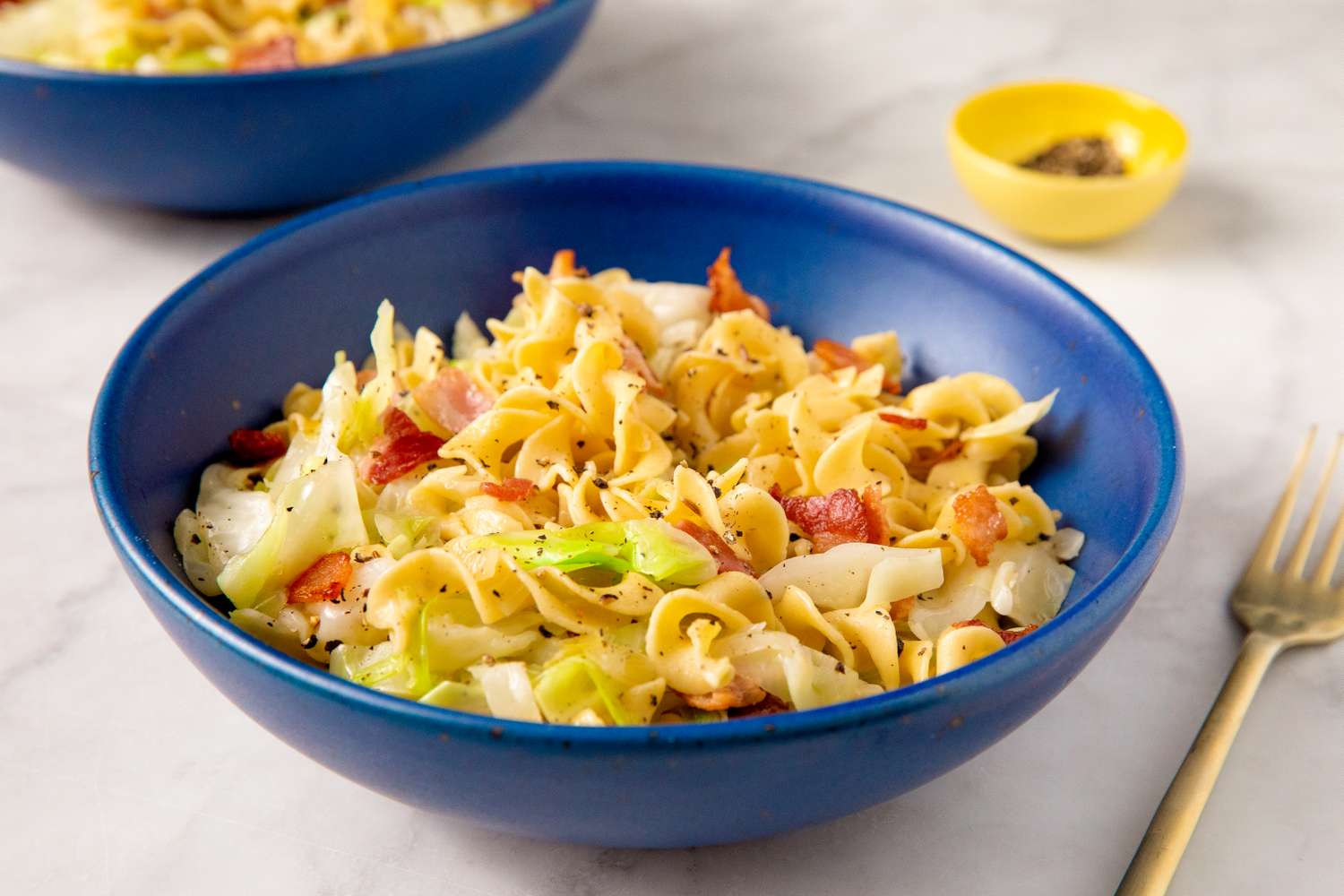 A bowl of haluski noodles with cabbage and bacon placed on a table with a golden fork and a small bowl of seasoning in the background