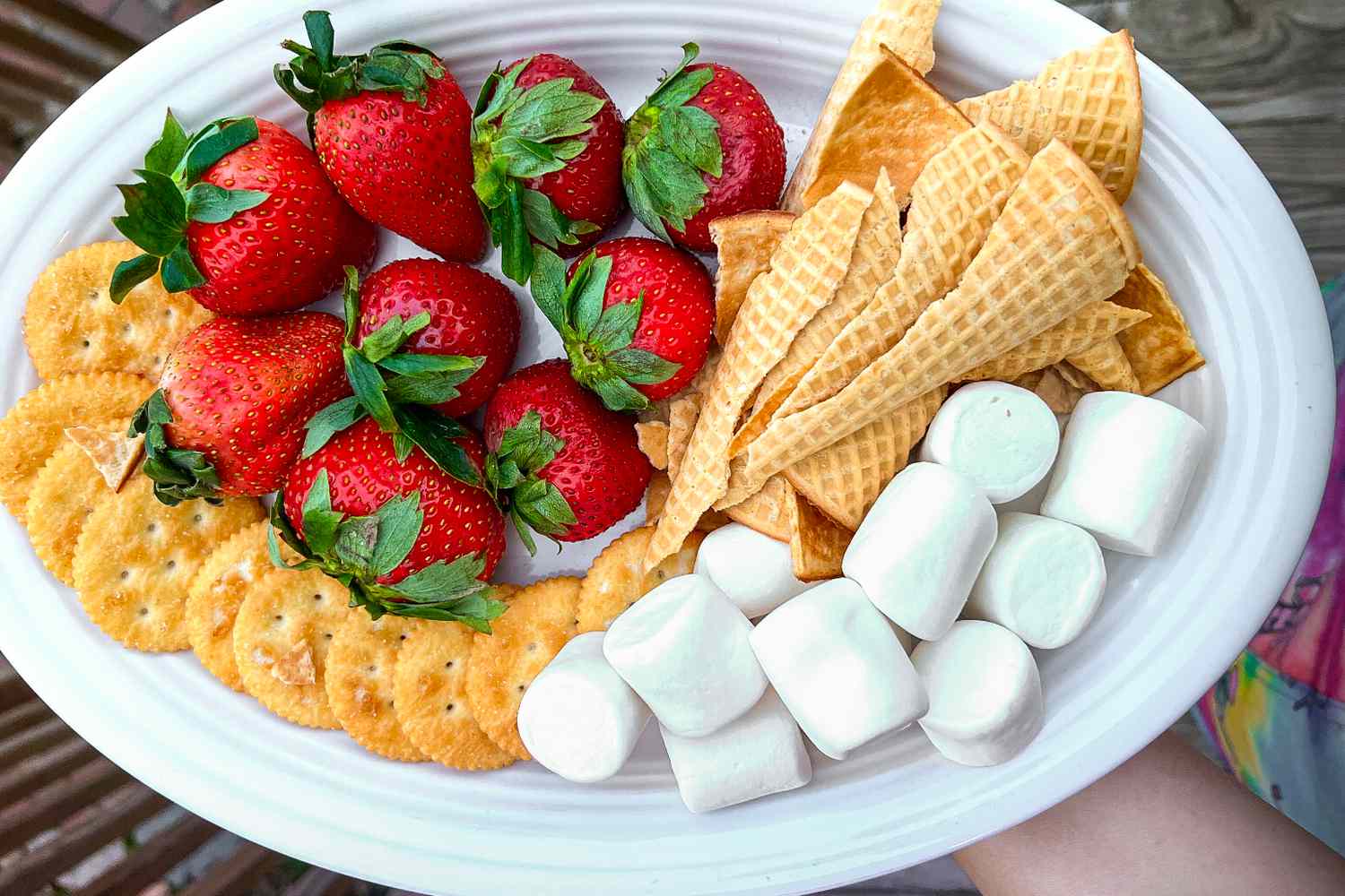 Overhead view of a white plate of strawberries, crackers, marshmallows and waffle cone pieces for dipping in cheese fondue