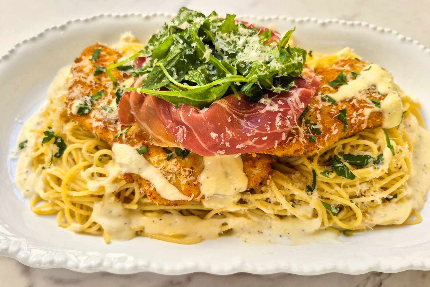 A plated dish featuring pasta topped with breaded chicken creamy sauce prosciutto and arugula on a decorative white plate