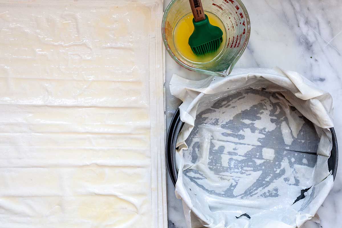 Sheets of phyllo are laid out next to a cake pan that has one sheet of phyllo laid inside and draped over the edge. A measuring glass of melted butter with a pastry brush inside.