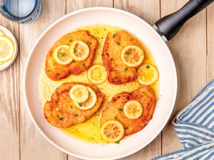 Italian butter chicken (petti di pollo al burro) in a skillet at a table with a plate of lemon slices, a glass of water, and a table napkin