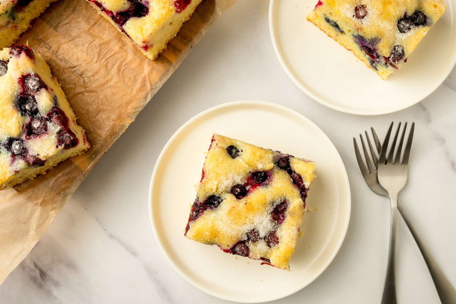 Squares of blueberry sheet cake on plates and parchment, with forks placed beside the plates