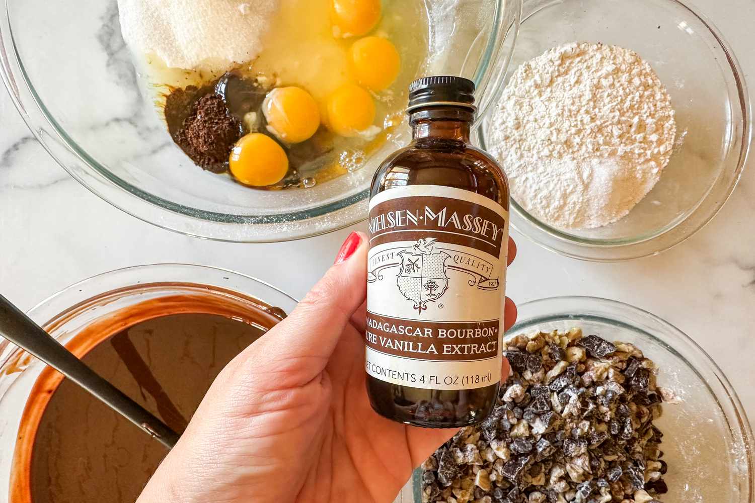 A hand holding a vanilla extract bottle with bowls of baking ingredients in the backgrounds