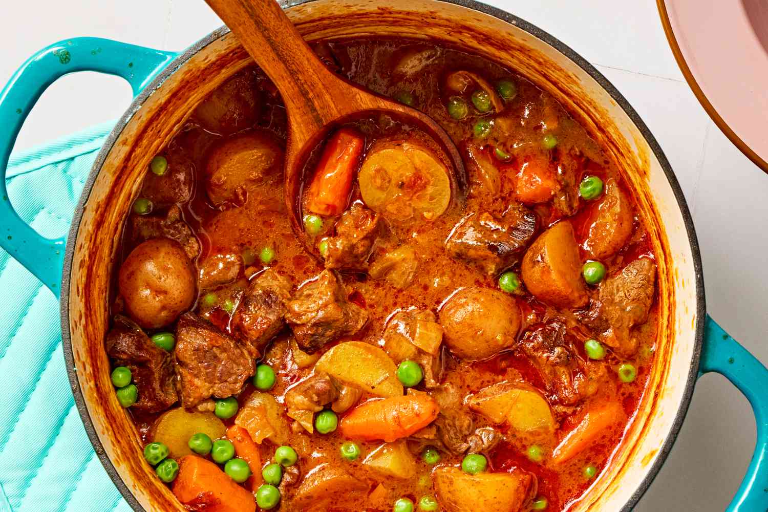 Overhead closeup view of a teal colored dutch oven of beef stew and a wooden spoon all on a tile countertop next to a pink bowl and blue hotpad