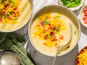 Overhead view of two bowls of potato soup topped with bacon, shredded cheese and green onions next to a bowls of bacon, green onions and shredded cheese