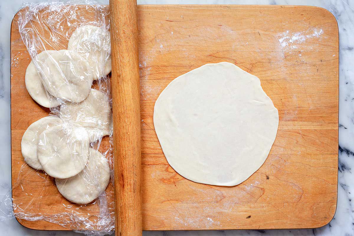 Small disks of dough to make flour tortillas are on a wooden cutting board. A rolled out tortilla is to the right on the cutting board and a rolling pin separates the disks from the finished tortilla.