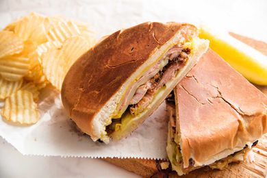 Two halves of a traditional cuban sandwich are resting on parchment paper and a wooden cutting board. Ridged potato chips and a pickle are in the background.
