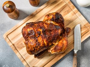 A whole roasted chicken on a wooden cutting board with a knife and condiment containers nearby