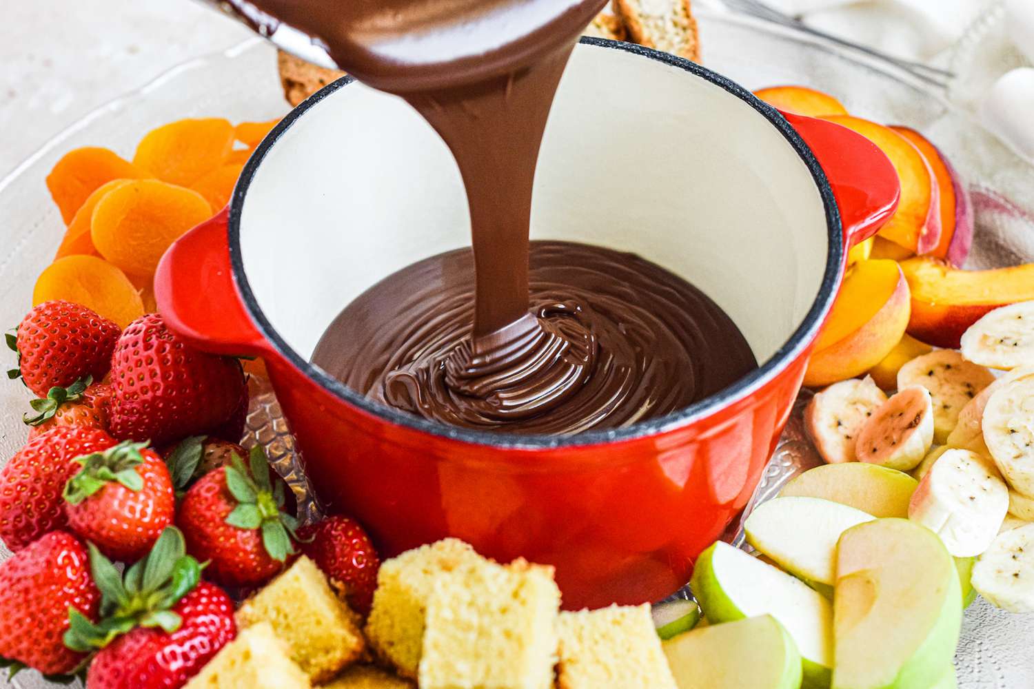 Pouring chocolate into a fondue pot surrounded by fruit.