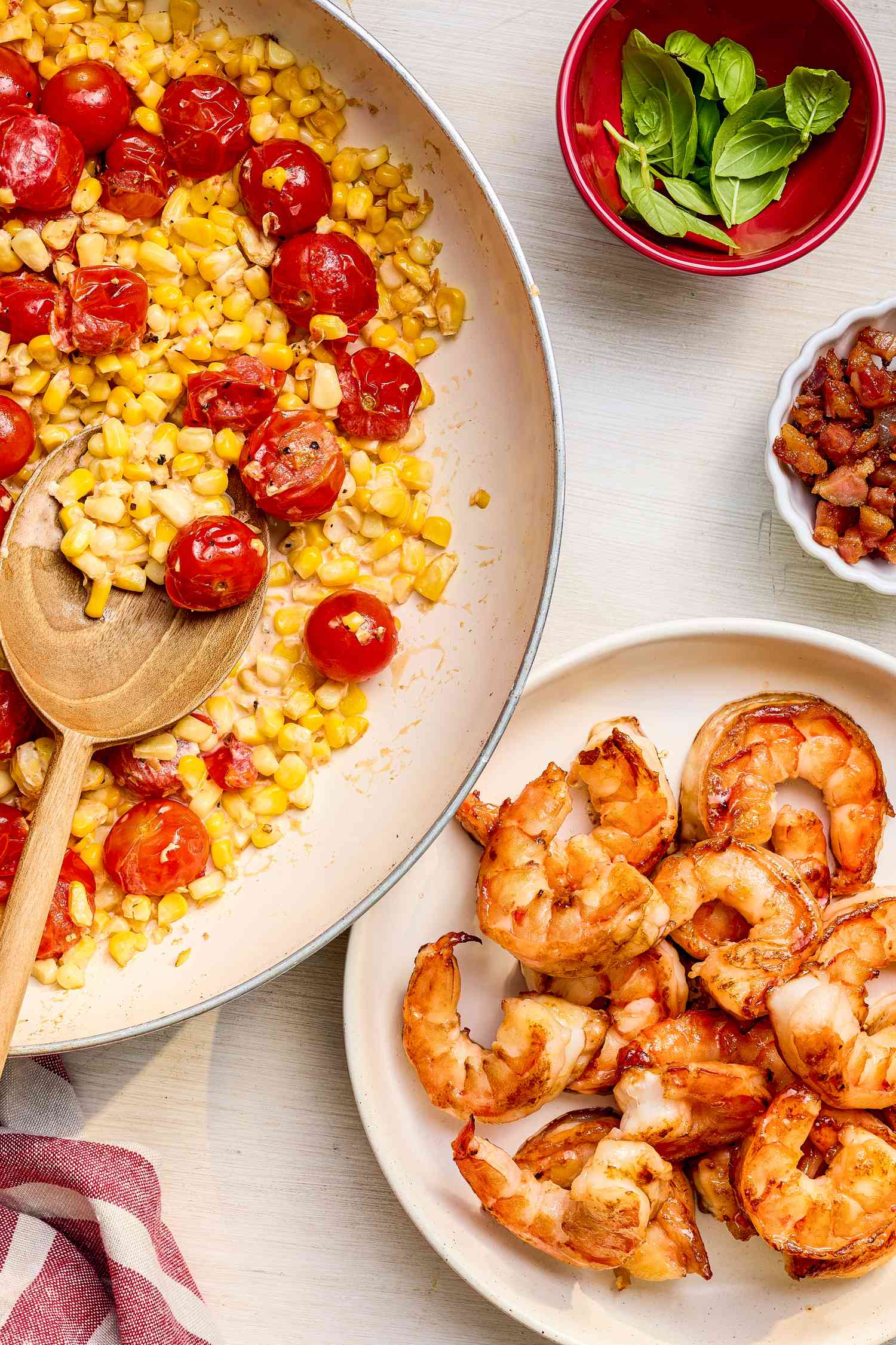 Skillet with roasted tomatoes and corn in a cream sauce next to a plate of stir-fried shrimp, a small bowl of pancetta, and a small bowl of basil leaves, all sitting on the counter