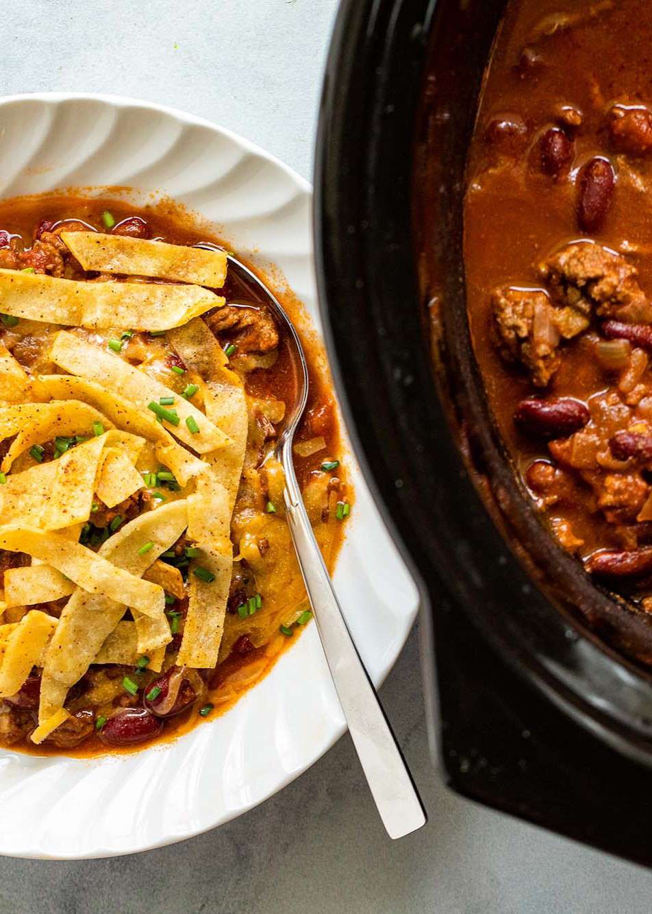 A bowl of beef and bean chili that's garnished with fried tortilla strips and sitting next to a slow cooker