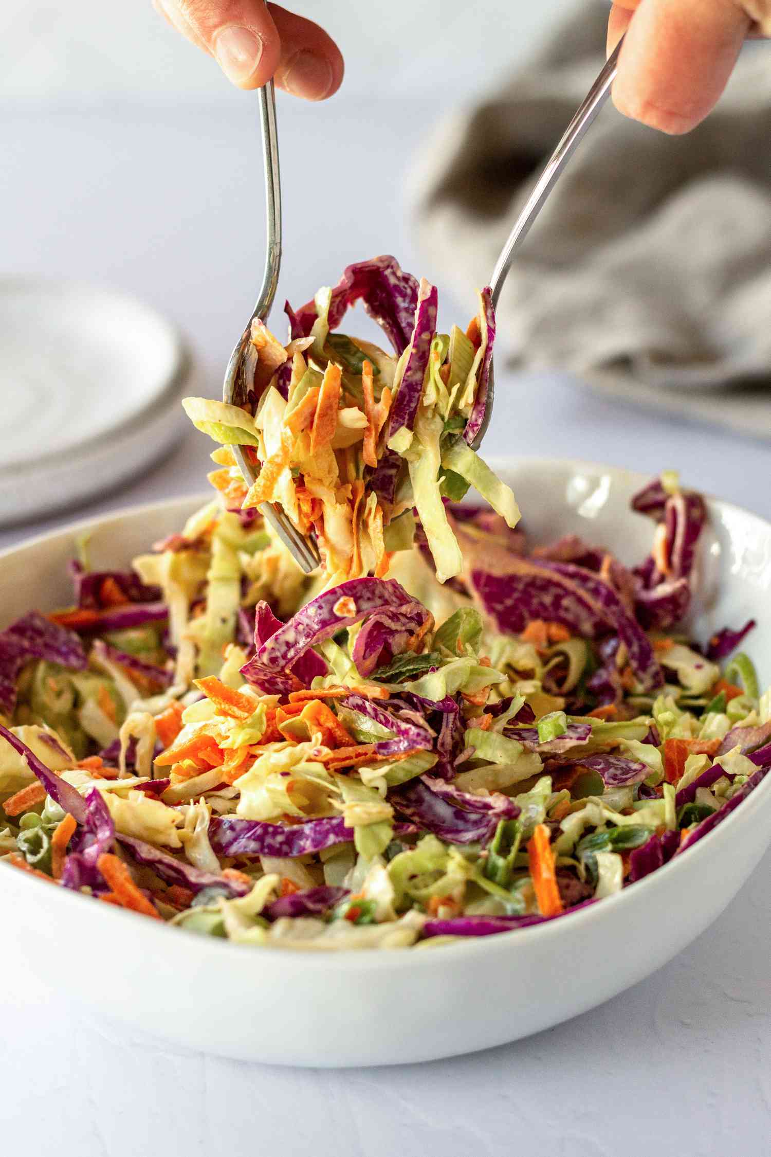 Forks lifting bright cabbage coleslaw out of a white bowl.