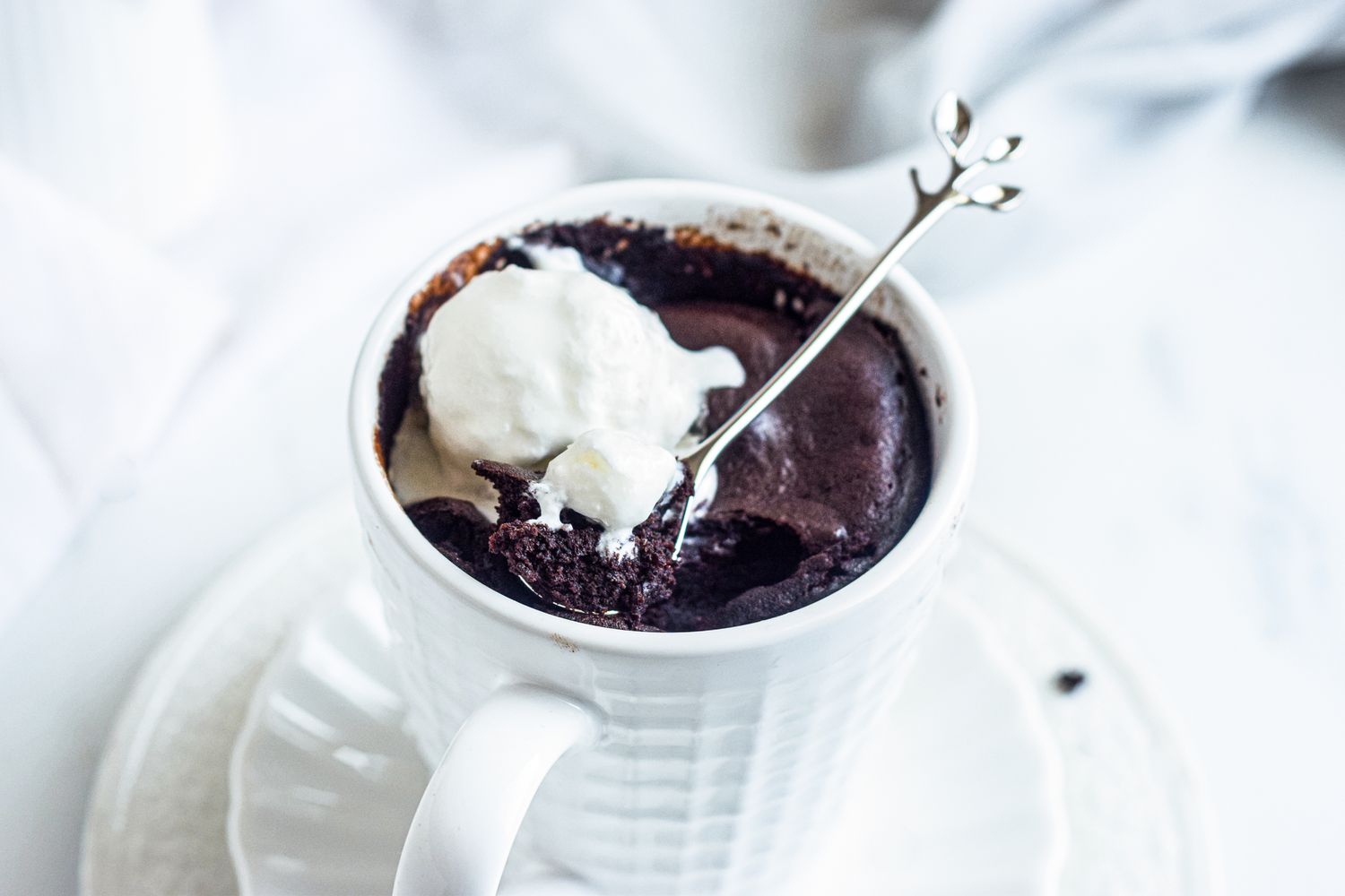 Side view of a brownie in a white mug and topped with ice cream and with a stir spoon inside.