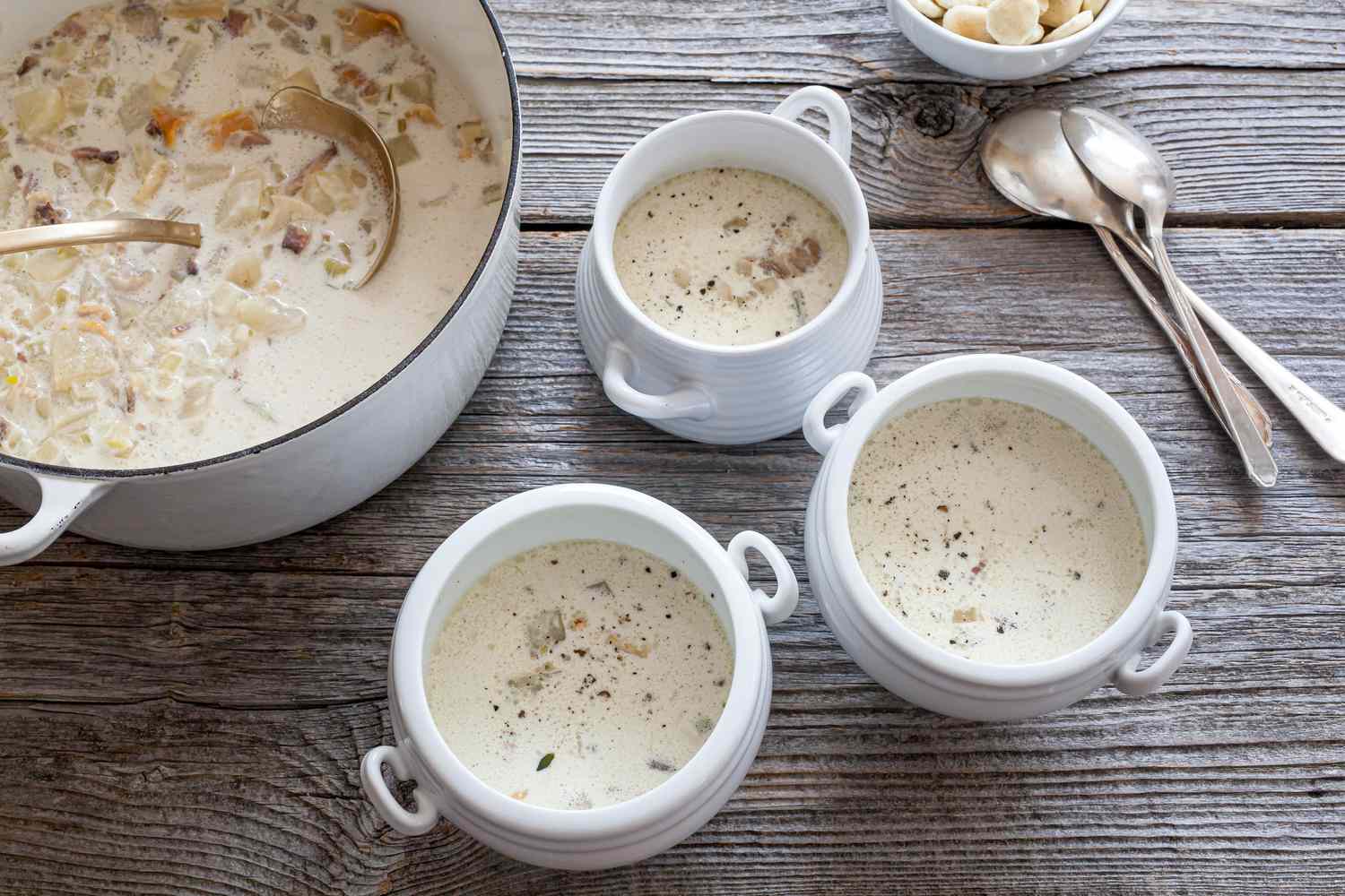 Classic New England Chowder in a Dutch Oven and in Individual Soup Bowls with Handles Next to a Couple of Spoons