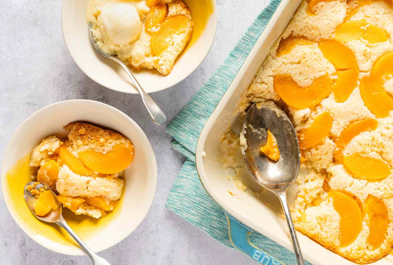 A peach cobbler in a baking dish beside two servings in bowls with spoons