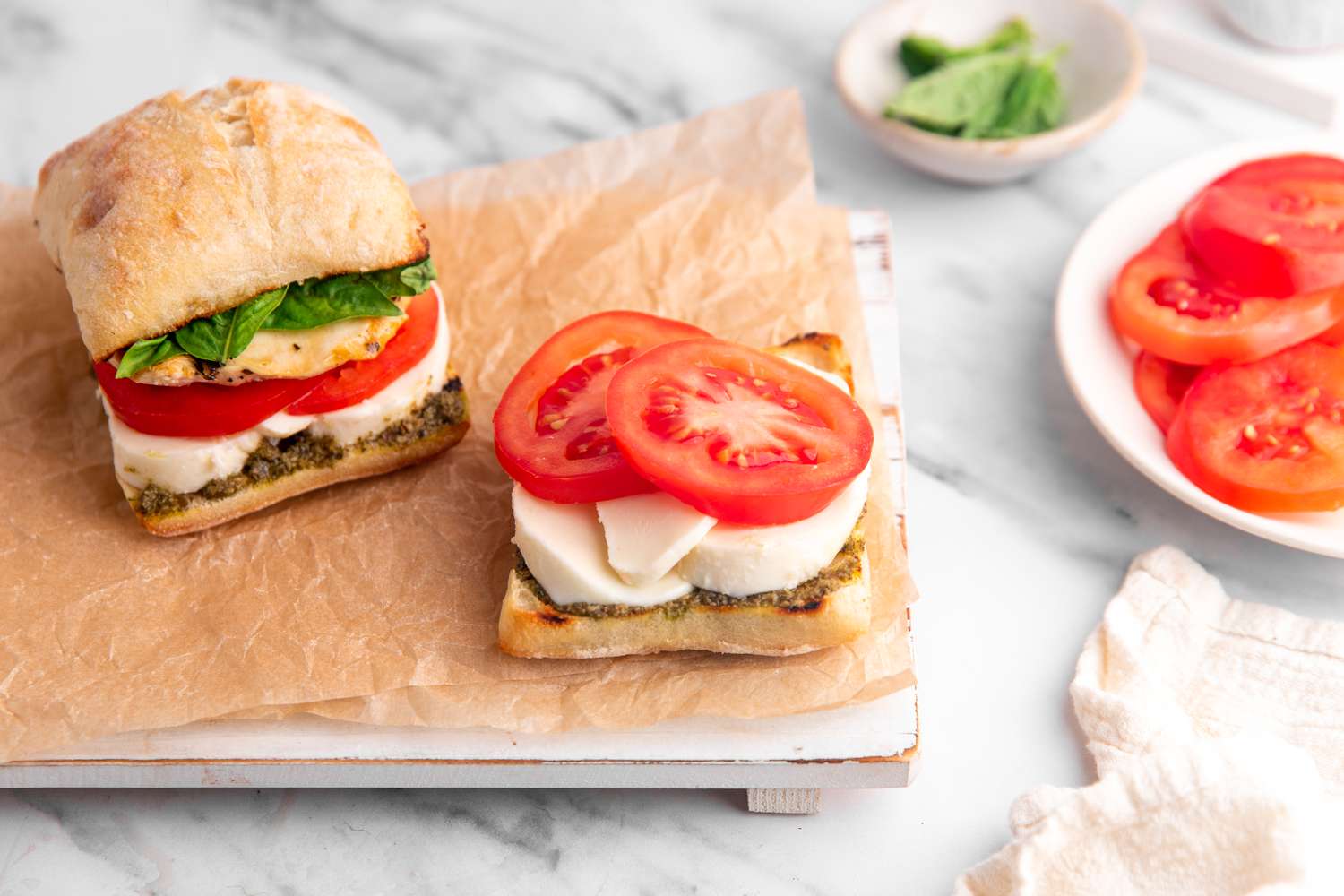 Grilled Chicken Caprese Sandwich Assembled on a Parchment Lined Tray (Another Assembled Sandwich Next to a Slice of Ciabatta With Pesto, Mozzarella, and Sliced Tomatoes), and on the Counter Next to the Tray, a Plates With More Ingredients 