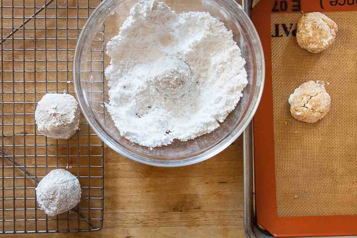 Two Mexican Wedding Cookies covered in powdered sugar on a wire rack, one in a bowl of powered sugar, and two on a baking sheet waiting to get covered in the powered sugar