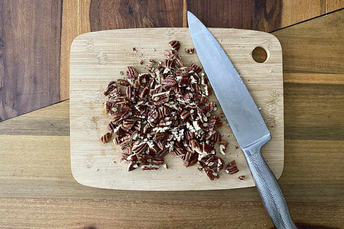 A cutting board with chopped pecans and a chef knife to make a Fall Vegetable Salad with Bacon and Maple Dijon Vinaigrette.